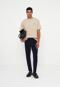 Beige lyhythihainen t-paita, jossa on pieni printti, laivaston siniset slim-fit housut, musta nahkakassi ja kengät, vaaleaa valkoista taustaa vasten.