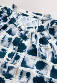 Bluzka z krótkim rękawem w abstrakcyjny wzór w kolorze granatowym i białym, wykonana z miękkiego materiału, z marszczonym dekoltem i metką wewnątrz.