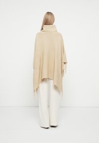 Poncho de malha bege com colarinho alto e textura canelada, apresentando bordas com franjas. Combinado com calças brancas de perna larga.