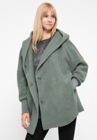 Grüne, fleecegefütterte Jacke mit lockerer Passform, großer Kapuze, zwei-Knopf-Verschluss und zwei Seitentaschen. Angenehme Textur, gemütliches Design.