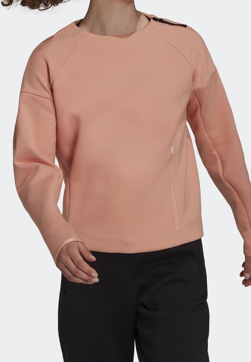 Femme portant un sweat-shirt pêche à manches longues avec zips aux épaules et pantalon noir, les bras légèrement pliés en mouvement de marche.