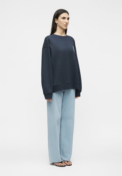 Femme debout, portant un sweat-shirt oversize bleu marine foncé, un jean large bleu clair et des talons ouverts noirs sur un fond clair uni.