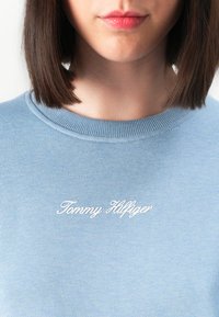 Lähikuva henkilöstä, joka käyttää vaaleansinistä hupparia, jossa on valkoisella käsinkirjoitettuna "Tommy Hilfiger" rintaan brodeerattu.