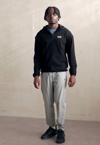 Musta vetoketjuhuppari logolla, harmaat kapeat housut ja mustat lenkkitossut; sileä kangas ja rento istuvuus. Malli seisoo neutraalissa taustassa.