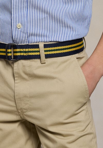 Osoba nosząca beżowe spodnie z paskiem z materiału w niebieskie, żółte i zielone paski oraz włożoną w spodnie niebiesko-białą koszulę w paski.