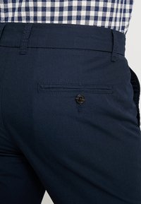 Pantaloni blu navy con tasca a filetto sul retro con bottone, indossati con una camicia a quadri blu e bianca infilata dentro.