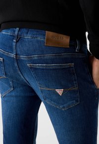 Jeansy denimowe w ciemnym niebieskim kolorze z skórzaną naszywką z logo "GUESS". Posiadają przeszywane kieszenie oraz kontrastujące przeszycia wzdłuż szwów.