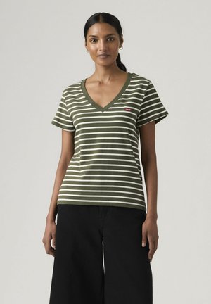 Kobieta o ciemnych włosach związanych w niski kucyk, ubrana w zielono-biały koszulka w paski z dekoltem w serek i czarne szerokie spodnie, stojąca na jednolitym tle.