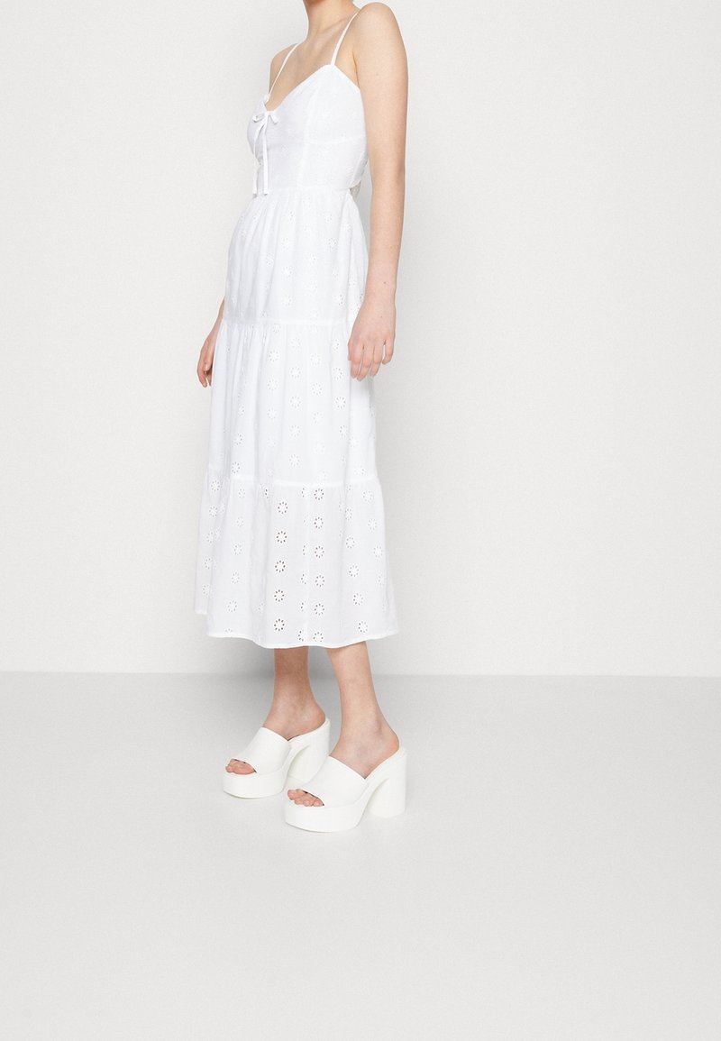 Femme portant une robe midi blanche sans manches avec des motifs œillets et des sandales à talons compensés blanches, debout contre un mur uni.