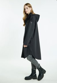 Impermeabile nero con cappuccio, caratterizzato da una vestibilità rilassata, bottoni frontali e tasche laterali, abbinato a jeans skinny grigi e stivali neri spessi.