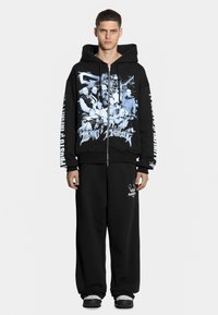 Czarna bluza z kapturem na zamek z niebieskim nadrukiem z przodu i na rękawie. W połączeniu z czarnymi, luźnymi spodniami dresowymi z białym detalem logo.