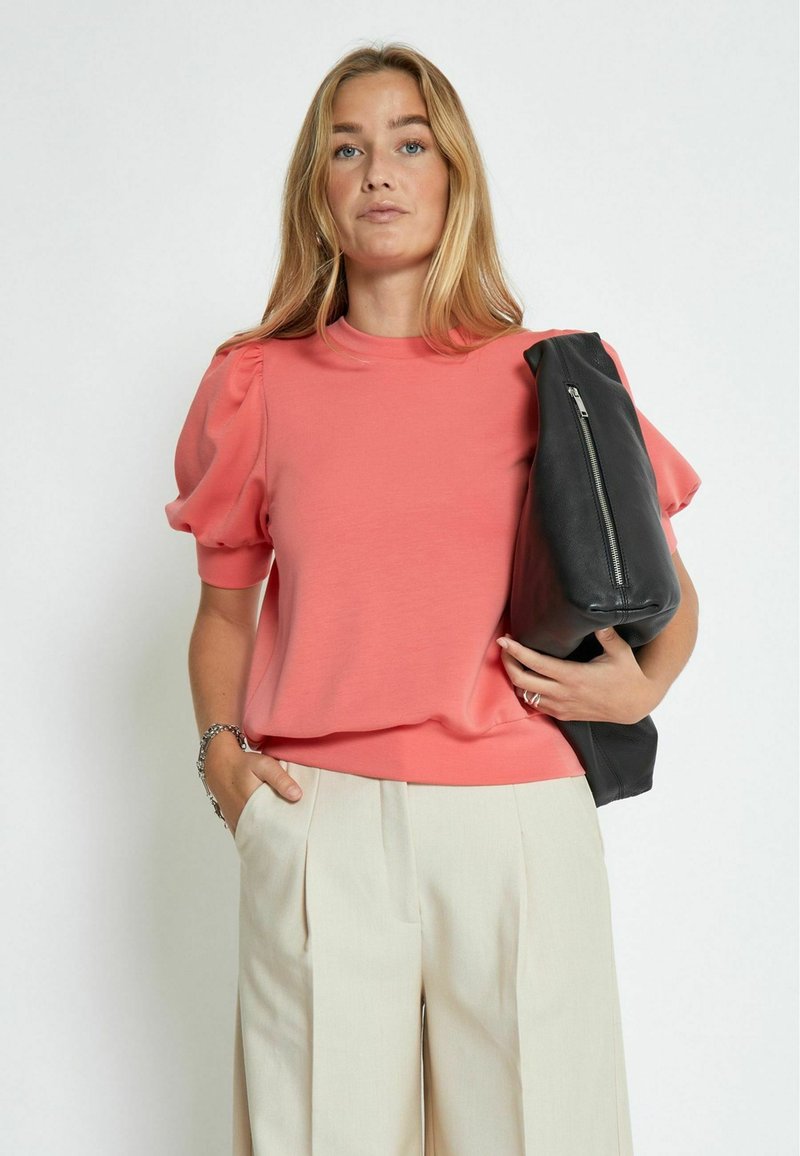 Top corto de color coral con mangas abullonadas, combinado con pantalones de tiro alto de color beige con pliegues y un bolso negro con cremallera. Textura de tela suave.