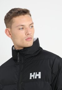 Juodas pūkinis švarkas su aukštu apykakle, užtrauktuku ir kontrastinga logo ant krūtinės. Lygi tekstūra ir siuvinėta struktūra.