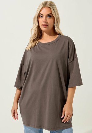 Blondynka w luźnej, krótkorękawowej brązowej koszulce i jasnoniebieskich dżinsach, stojąca na tle jednolitego jasnego tła.