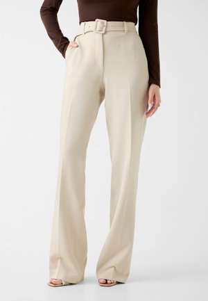 Pantalon beige taille haute à jambes larges avec ceinture assortie, porté avec un haut marron à manches longues et des talons ouverts beige.