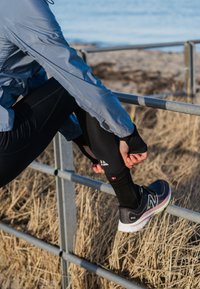 Atlet i en lyseblå jakke og sorte leggings, som strækker sig nær et metalrækværk, iført sorte sneakers med grønne detaljer.