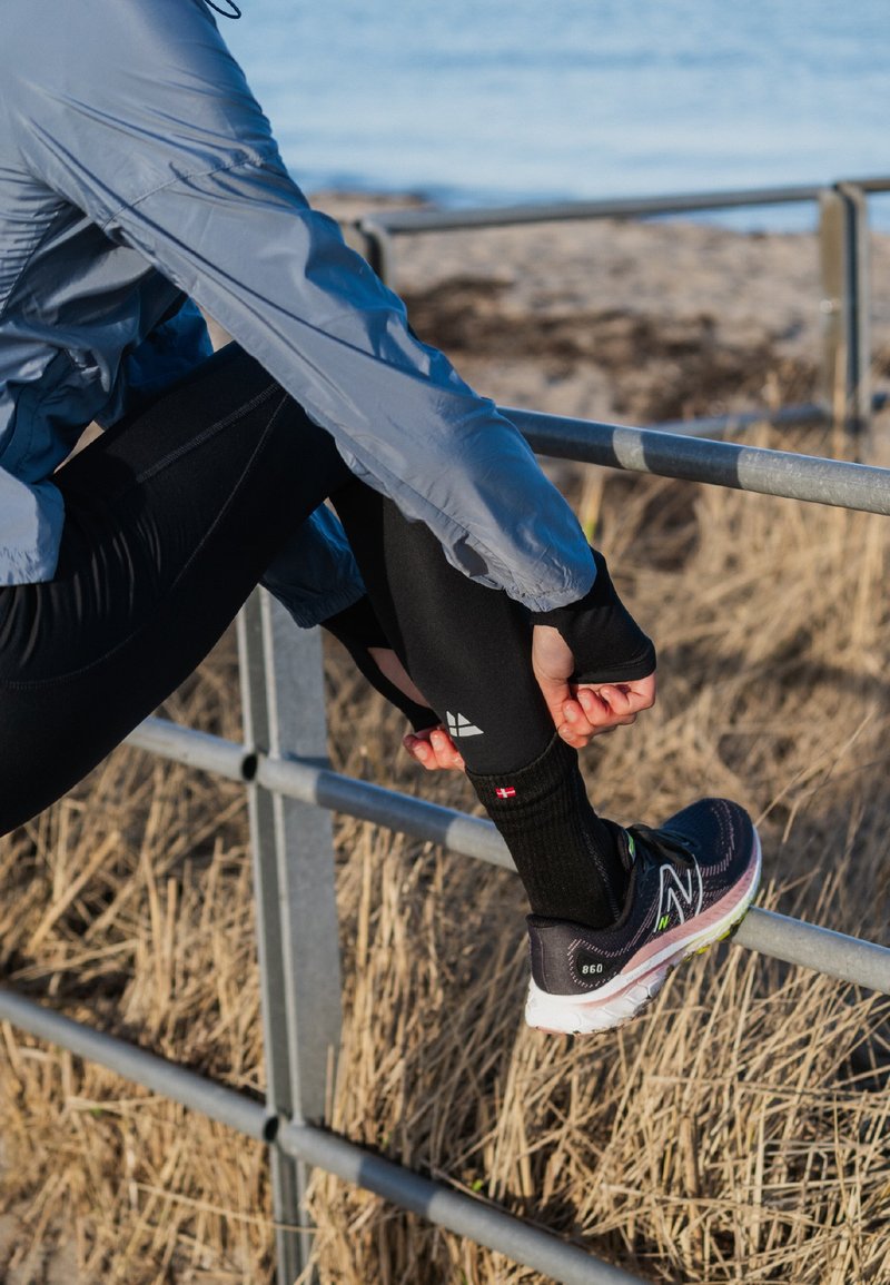 Atlet i en lyseblå jakke og sorte leggings, som strækker sig nær et metalrækværk, iført sorte sneakers med grønne detaljer.