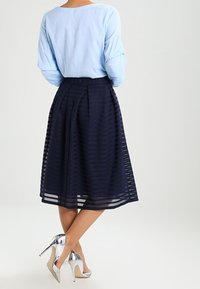 Žena v světle modré blůze s dlouhým rukávem, námořnicky modré plisované sukni po kolena a stříbrných vysokých podpatcích, stojící se zkříženýma nohama.