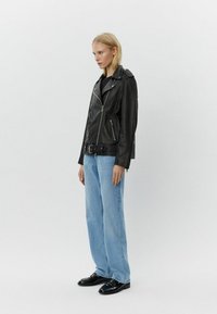 Chaqueta de moto de cuero negro con cremallera asimétrica, bolsillos laterales y cinturón en la cintura, combinada con jeans rectos de color azul claro y zapatos negros.