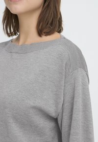 Naine kannab heledat halli kudumisjacket, millel on ribidega detailid õlgadel ja kaeluses, raamistatud pruuni õlgadeni ulatuva juuksevärviga.