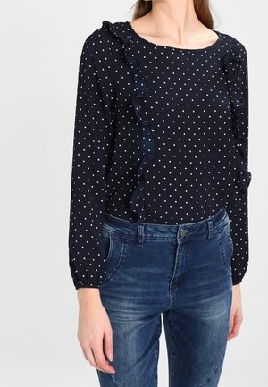 Femme portant un chemisier bleu marine à manches longues avec des pois blancs et des volants, associé à un jean bleu en denim.