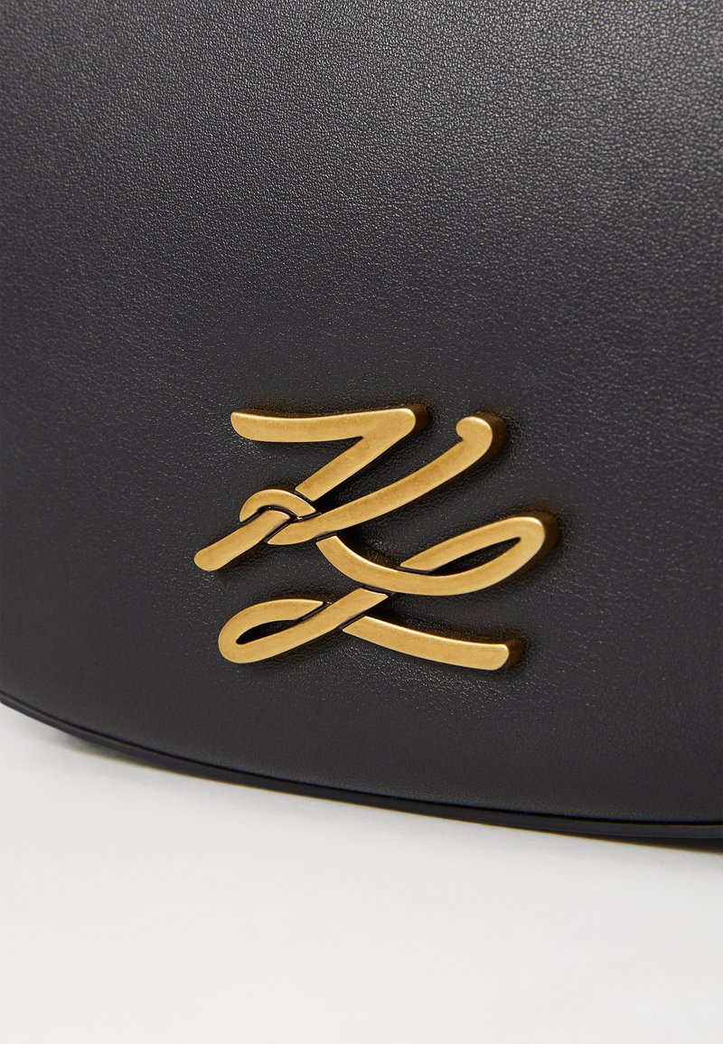Schwarze, strukturierte Handtasche mit einem auffälligen goldenen geschwungenen Logo. Das Material wirkt glatt und hat eine klare Form mit minimalen Details.