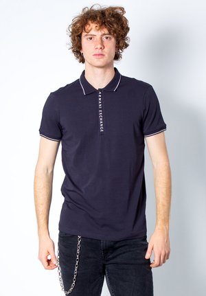 Polo navy con ribetes blancos y logo "ARMANI EXCHANGE" en la tapeta. Corte ajustado, mangas cortas, combinado con jeans oscuros y accesorio de cadena.