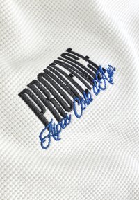 Tessuto bianco strutturato con logo ricamato in nero e blu, caratterizzato da un design ondulato e lettere distintive.