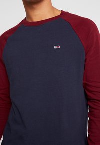 Pitkähihaisessa paidassa on tummansininen vartalo ja viininpunaiset raglan-hihnat, ja se on valmistettu puuvillasta. Rinnassa on pieni logo. Pehmeä rakenne.
