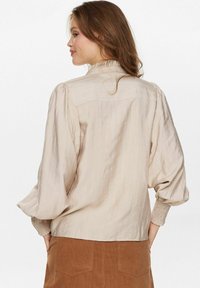 Beige, textúrájú blúz ráncos gallérral és puffos ujjal, függőleges csíkokkal és elasztikus mandzsettával, barna szoknyával párosítva.