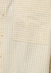 Chemise à carreaux beige clair avec une poche avant et des boutons, avec un petit logo brodé "R" sur la poche.
