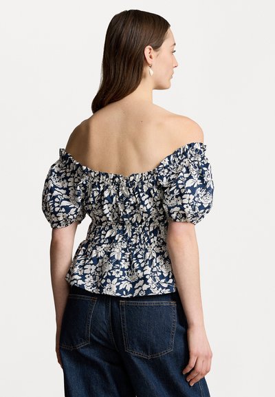 Jūras spēku ziedu blūze bez plecu siksnām ar pūkaino piedurkņu, savilktu aizmuguri un pepluma apakšu. Mīksts kokvilnas audums ar baltām ziedu apdrukām. Saskaņota ar tumšām denima biksēm.