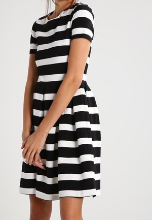 Femme portant une robe aux manches courtes, mi-longue, à rayures horizontales noires et blanches, debout les mains jointes devant elle.