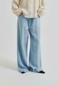 Svetlo modri široki kavbojki iz denima. Imajo sproščen kroj, visok pas in sproščen stil. V kombinaciji s kremno, teksturirano bluzo.