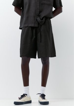 Osoba ubrana w czarną koszulę zapinaną na guziki i szorty, białe skarpetki oraz beżowe sneakersy z czarnymi podeszwami i sznurowadłami, stojąca na jednolitym tle.