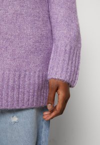 Main gauche d'une personne portant une manche de pull tricoté lavande et un jean en denim bleu clair usé, sur un fond uni.