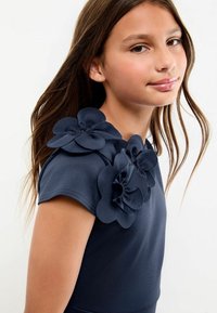 Top bleumarin cu mâneci scurte, decorat cu trei flori din țesătură la umăr. Textură netedă și design ajustat.