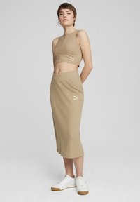 Beige ribbet crop top med PUMA-logo, sat sammen med en matchende blyantskirt. Modellen bærer hvide sneakers. Glat tekstur; tætsiddende silhuet.