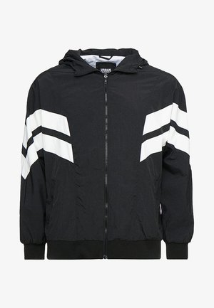 Veste noire avec capuche, fermeture éclair sur le devant et rayures blanches en diagonale sur les manches. Elle dispose de poignets élastiques et d'une coupe décontractée. Fabriquée en matériau léger.