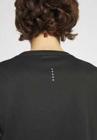 Schwarzes Sportshirt aus strukturiertem Stoff mit rundem Ausschnitt und einem weißen grafischen Design aus fünf Pluszeichen, das mittig im oberen Rückenbereich angebracht ist.