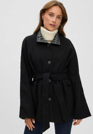 Un mannequin porte un manteau noir ceinturé à manches larges, avec un col à carreaux, un tour de cou en maille couleur crème et un jean foncé, sur un fond uni.
