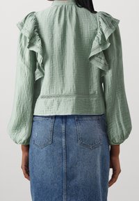 Blouse en tissu vert froissé avec des détails froncés et des accents perforés, associée à une jupe en denim avec poches arrière et un design droit.
