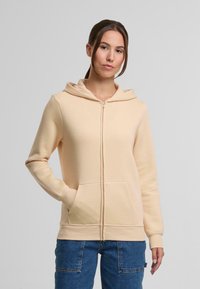 Cazadora beige con cremallera, bolsillo frontal, mangas largas y capucha. Hecha de una tela suave con una textura lisa y un diseño minimalista.