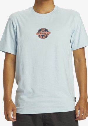 Maglietta a maniche corte azzurro chiaro con piccolo logo tondo Quiksilver raffigurante un globo sul petto, indossata da una persona.