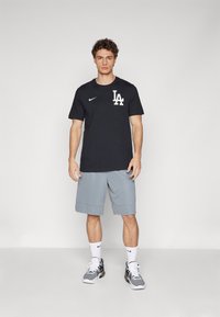 Black Nike t-shirt with white "LA" logo, paired with light gray athletic shorts. The model wears white Nike socks and gray sneakers.