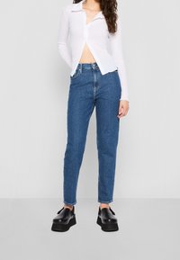 Vit långärmad skjorta med knappar och en kortad design, i kombination med blå högmidjade jeans och svarta platåskor. Släta texturer och rena linjer.