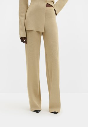 Person trägt beige weite Hosen und passendes langärmeliges Oberteil, steht mit einer Hand in der Hüfte, schwarze spitze Schuhe sichtbar.