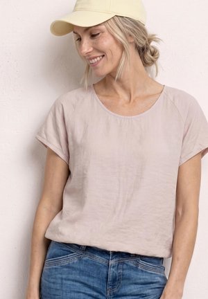 Vrouw met beige pet en lichtroze top met korte mouwen die lacht, staand tegen een bleke muur, draagt blauwe spijkerbroek.