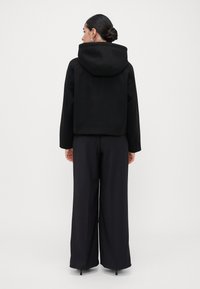 Vrouw met een bun in haar haar, gekleed in een zwarte cropped hoodie, wijde zwarte broek en hoge hakken, staande met haar rug naar de camera.
