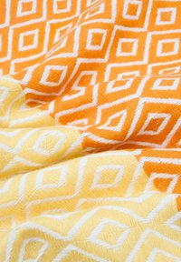 Bomuldsstof med et geometrisk mønster i orange og gult, med hvide kanter. Teksturen er vævet og let plyssede.